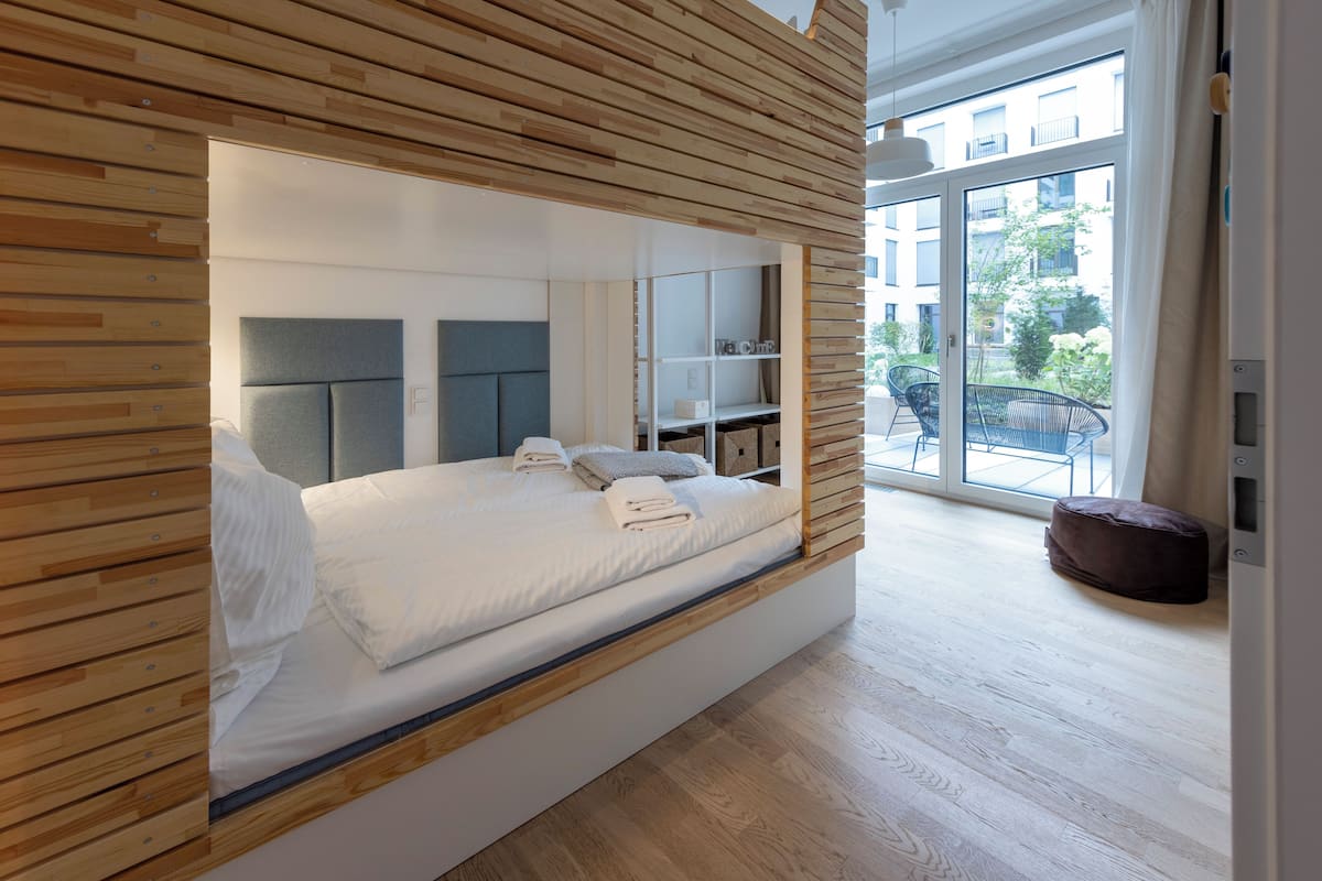 A modern bedroom with a built-in bed, wooden paneling, a large window, and a balcony with outdoor furniture.
