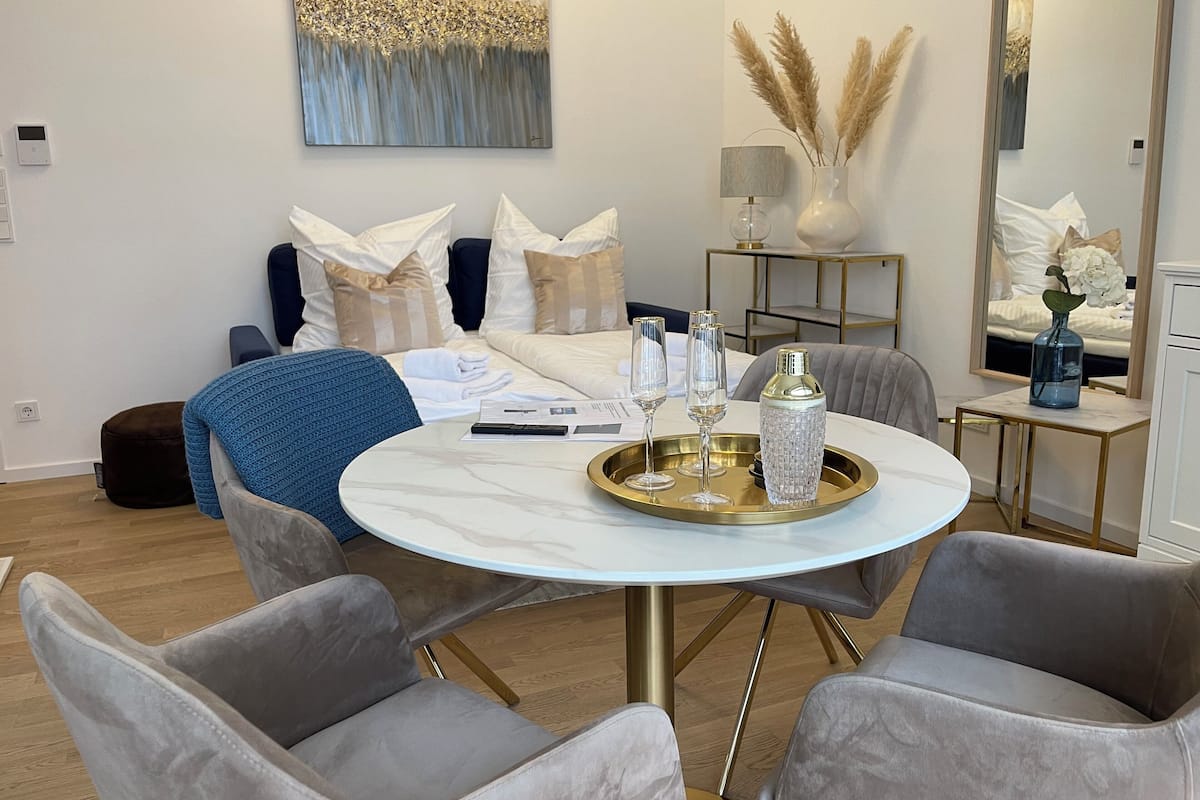 A modern hotel room with a bed, a round dining table with chairs, a bedside table with a lamp, and a large mirror.