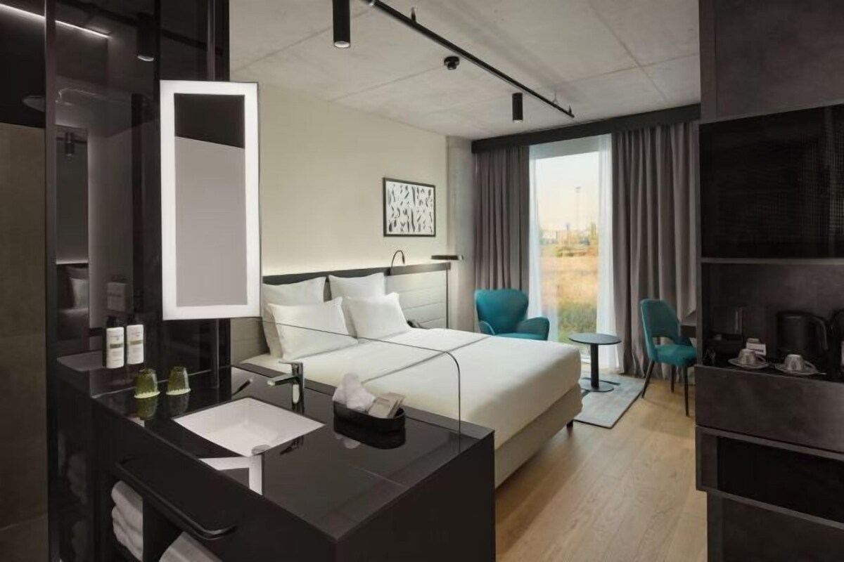 A modern hotel room with a large bed, a separate seating area, a small table, and a large window with curtains.