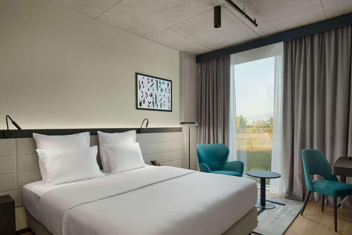 A hotel room with a large bed, two chairs, a small table, and a view of the outdoors.