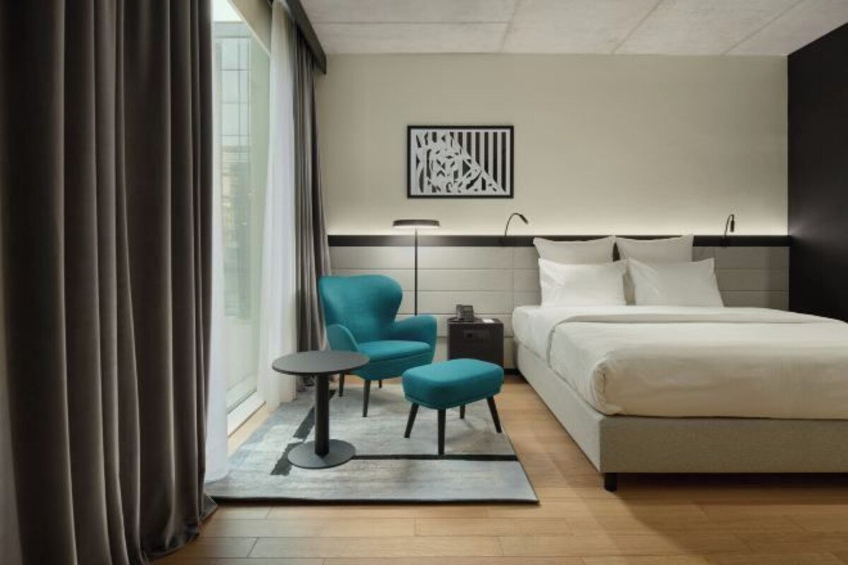 A modern hotel room with a large bed, a small table, two chairs, a nightstand, and a window with curtains.
