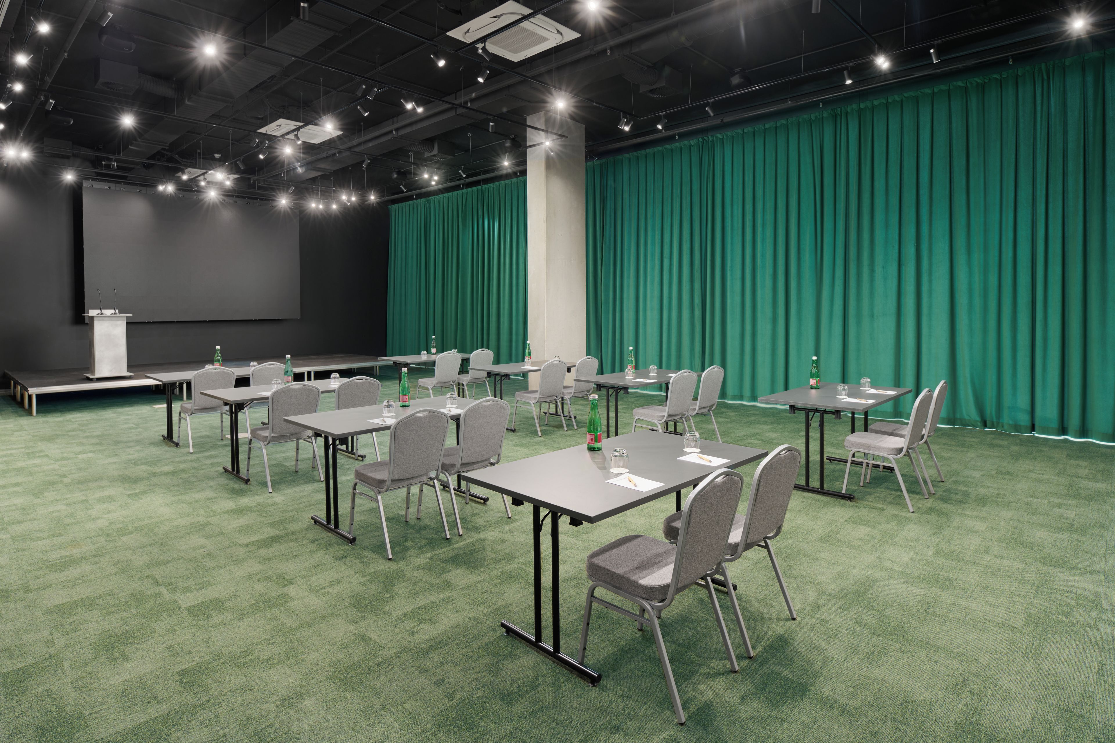 Una sala de conferencias con alfombra verde, sillas grises y mesas, un escenario con una pantalla y un telón de fondo verde.