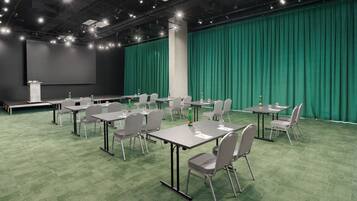 Una sala de conferencias con alfombra verde, sillas grises y mesas, un escenario con una pantalla y un telón de fondo verde.