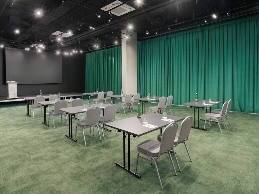 Una sala de conferencias con alfombra verde, sillas grises y mesas, un escenario con una pantalla y un telón de fondo verde.