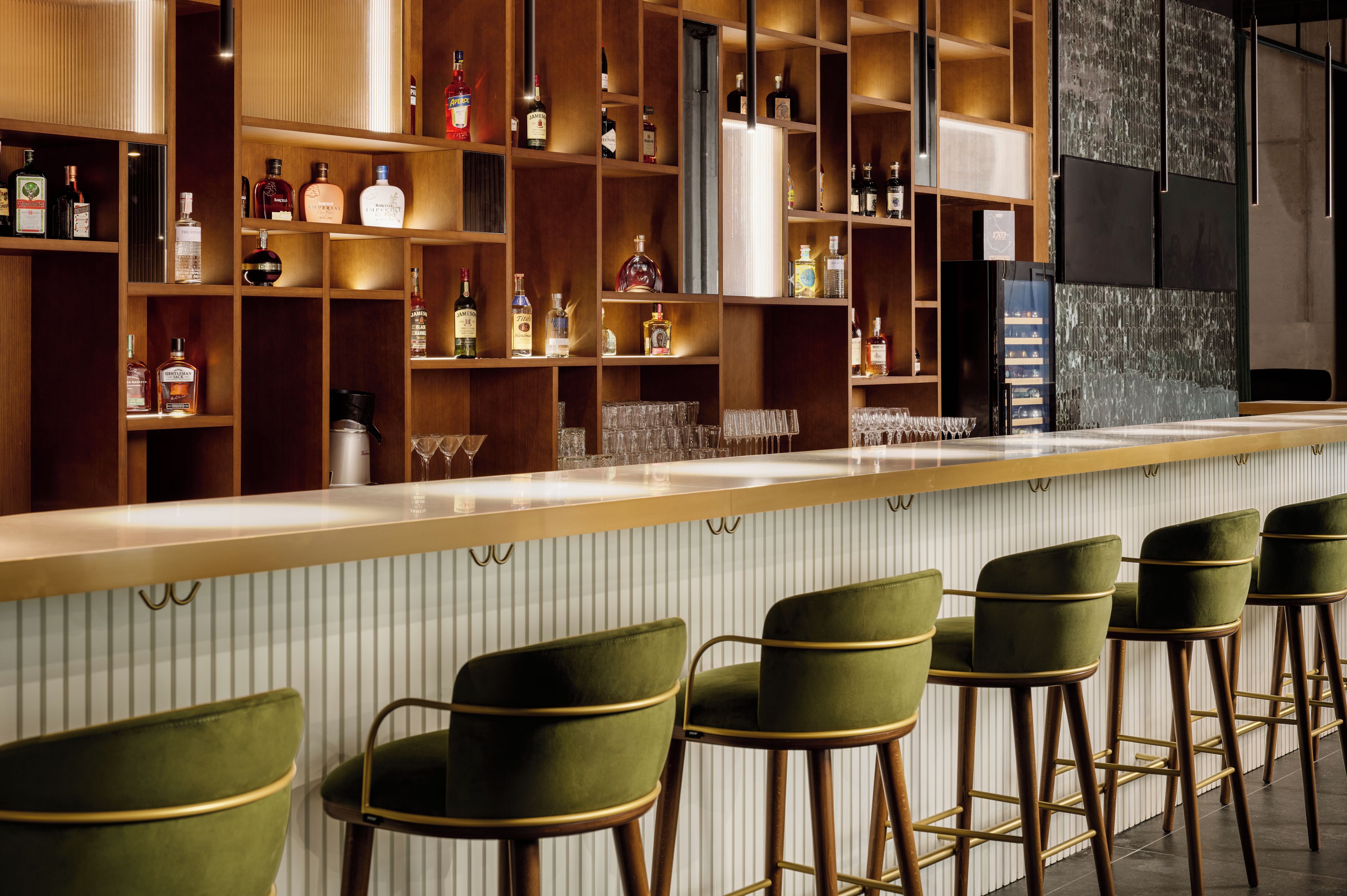Una zona de bar con estanterías de madera llenas de botellas y vasos, y taburetes de bar verdes.