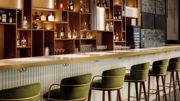 Una zona de bar con estanterías de madera llenas de botellas y vasos, y taburetes de bar verdes.