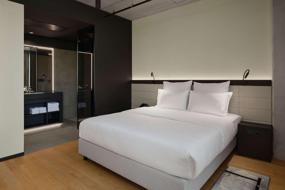 A modern hotel room with a large bed, a bedside table, a nightstand, a built-in wardrobe, and a bathroom with a mirror and sink.