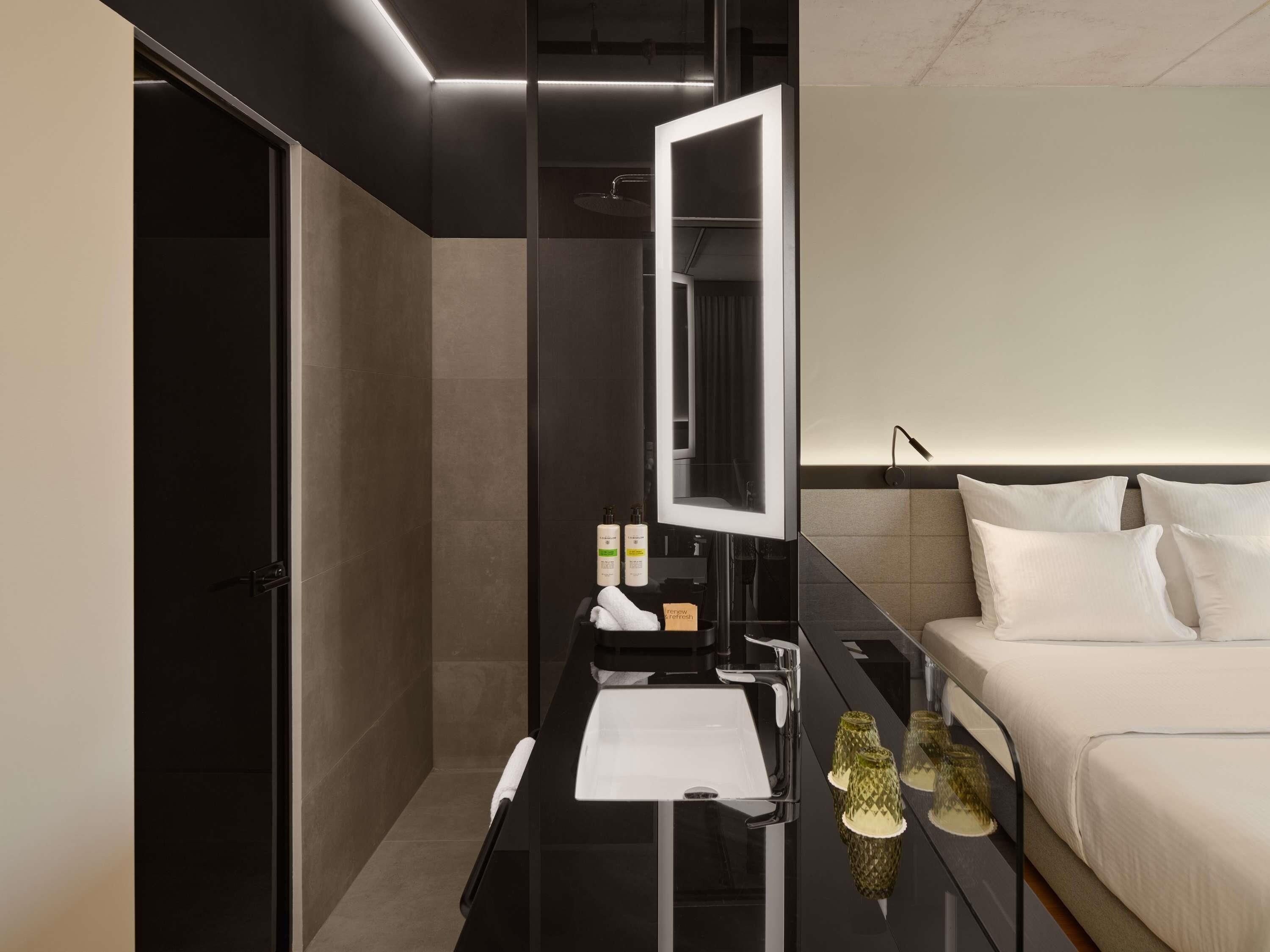 A modern hotel room with a large mirror, a bed, a bedside table with toiletries, and a nightstand with decorative items.