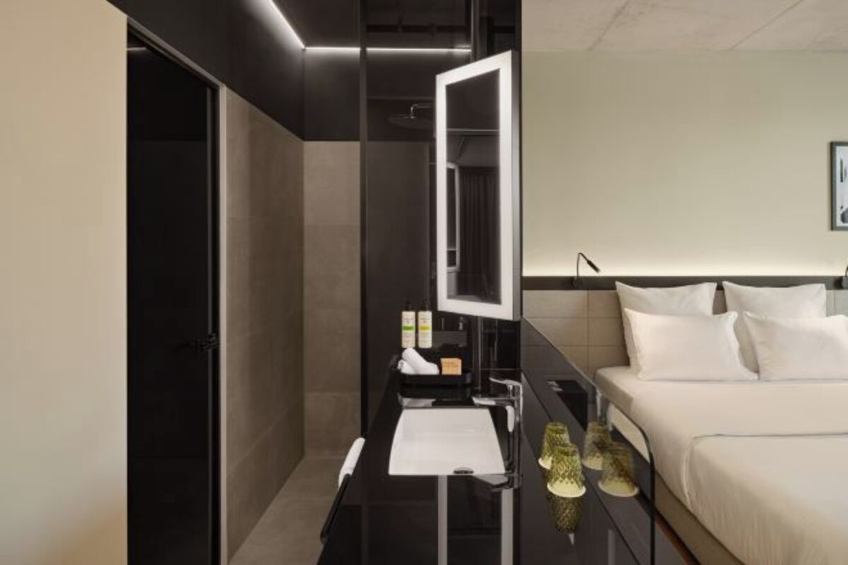A modern hotel room with a large bed, a bedside table with a mirror, and a small desk with a lamp.