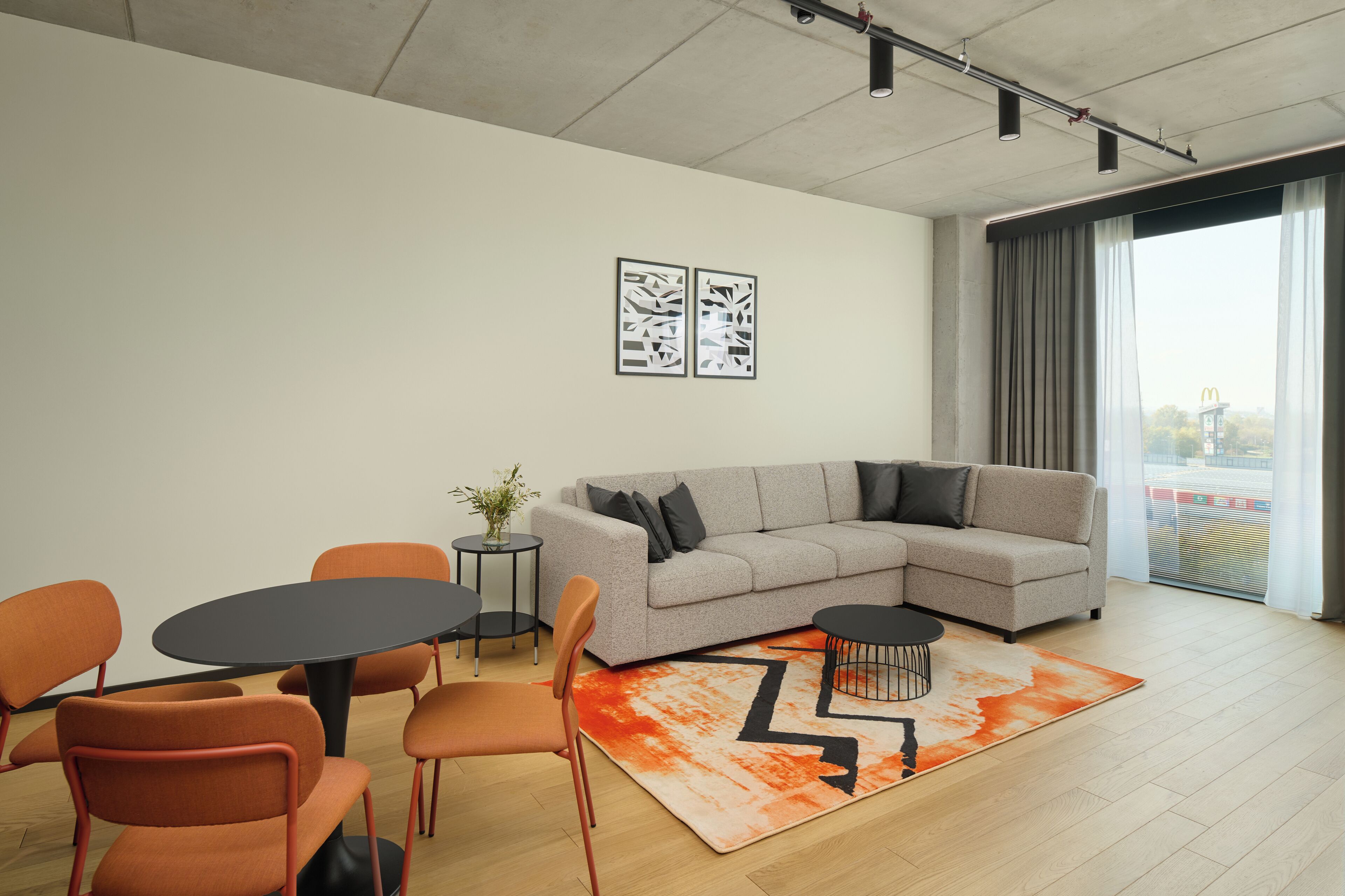 Una sala moderna con un sofá seccional gris, sillas naranjas, una mesa redonda y un amplio ventanal con cortinas.