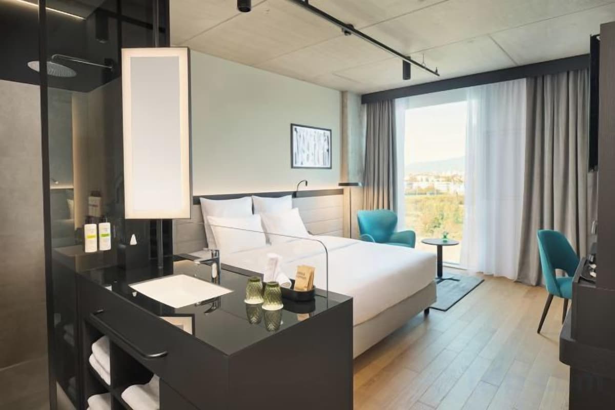 A hotel room with a bed, a desk with a mirror, a chair, and a view of the outdoors.