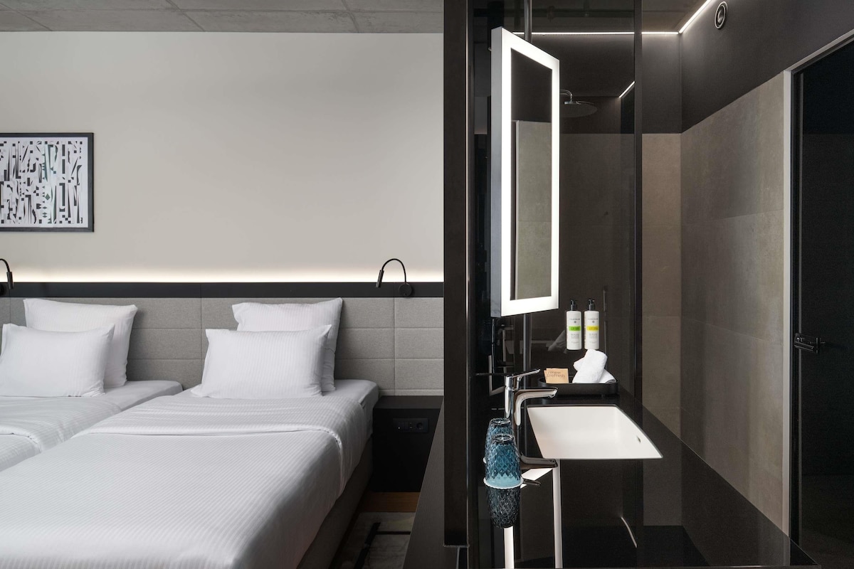 A modern hotel room with a large bed, a bedside table, a mirror, and a bathroom with a sink and shower.