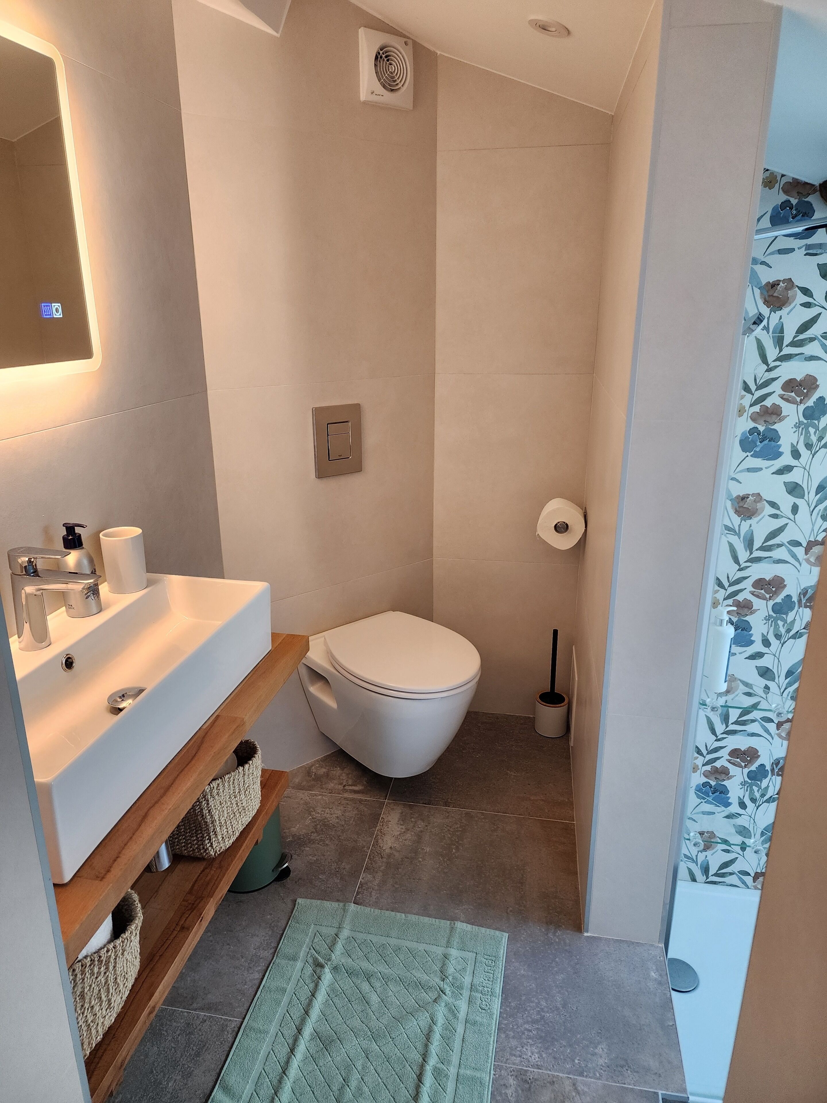 A bathroom with a white sink, toilet, and shower. There is a mirror above the sink, a towel rack, and a green bath mat.