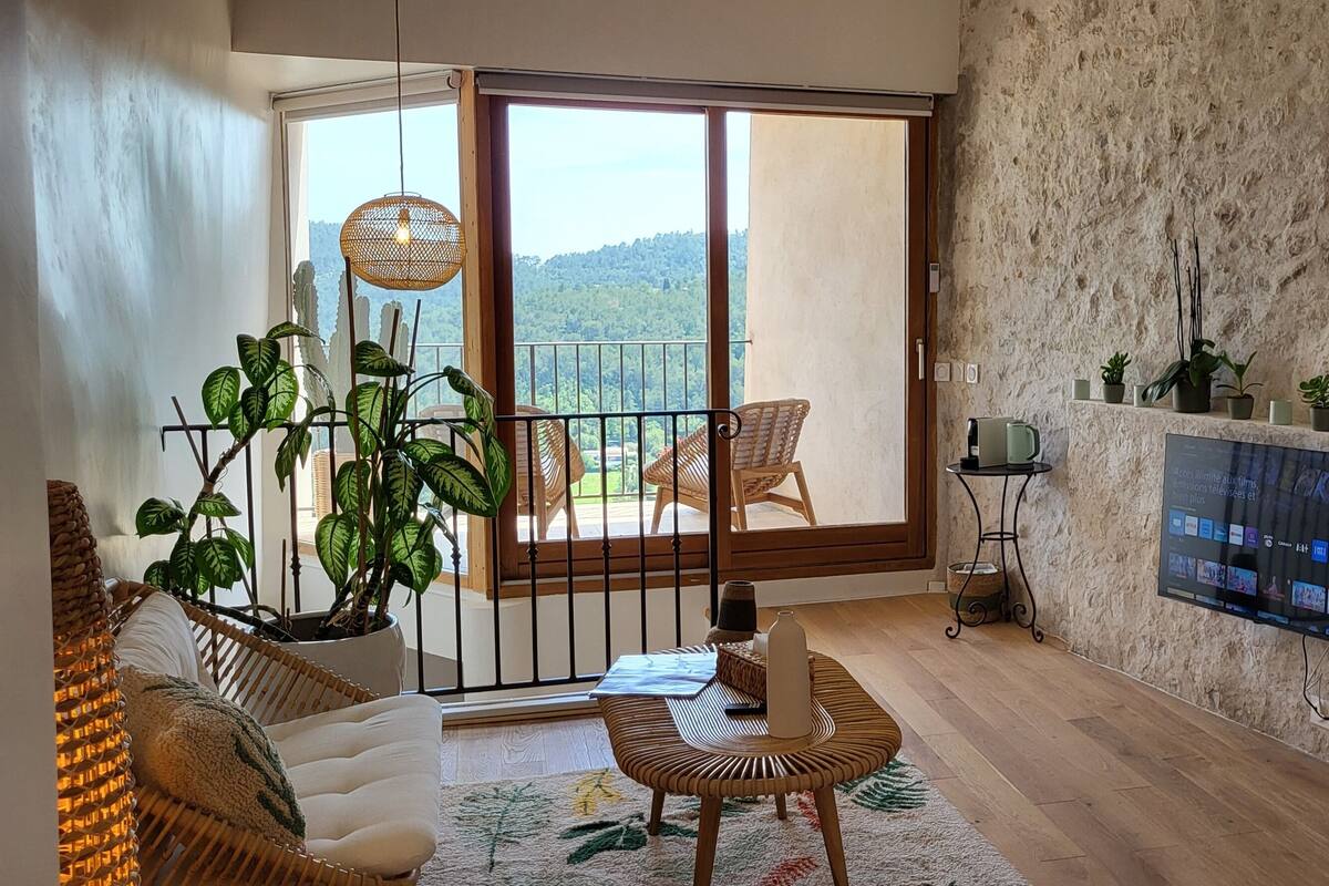 A living room with a large sliding glass door leading to a balcony, a mounted TV, and a variety of potted plants.
