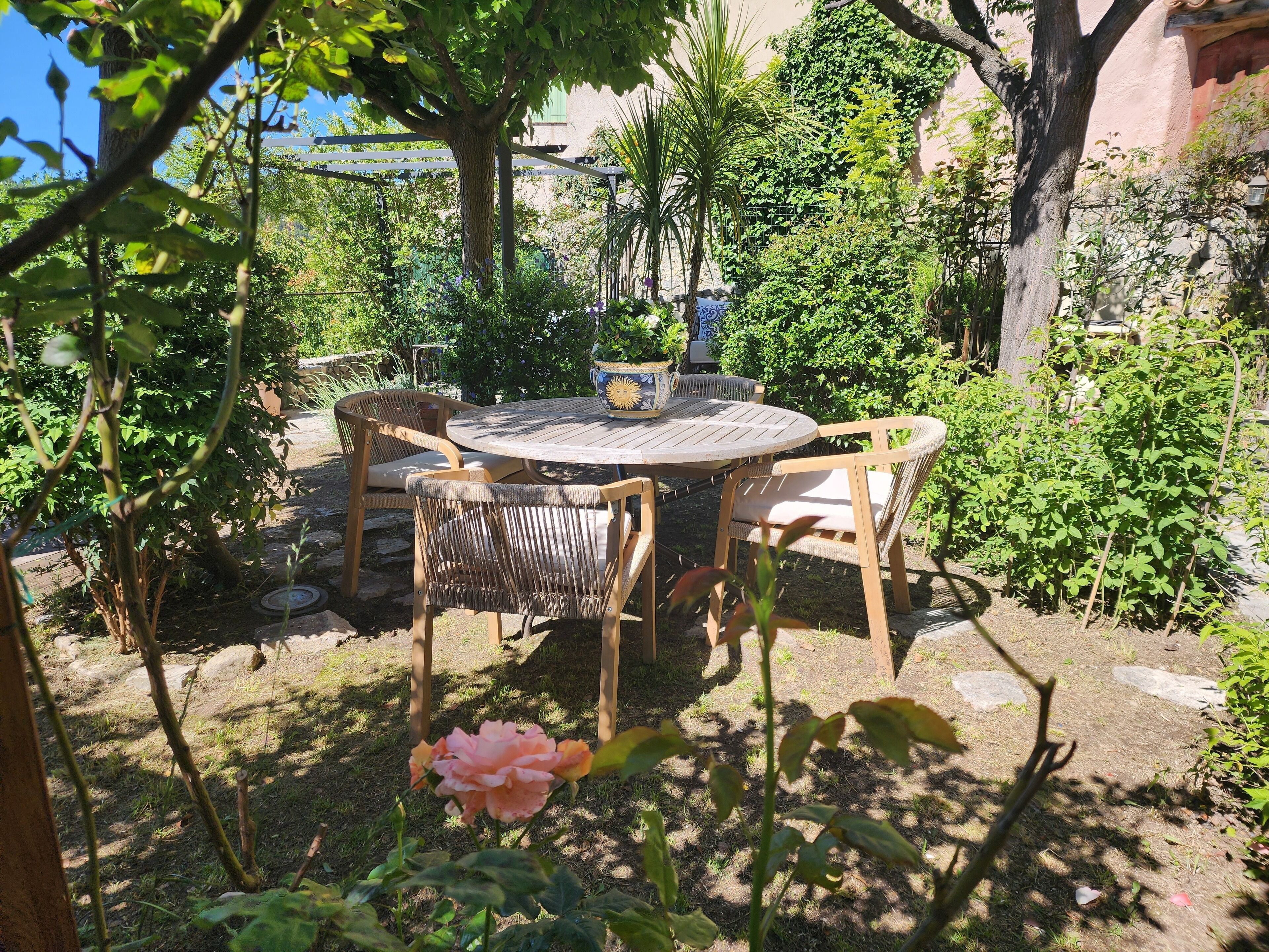 Une table ronde en bois, entourée de quatre chaises, et bordée d’une végétation luxuriante et d’une plante en fleurs.