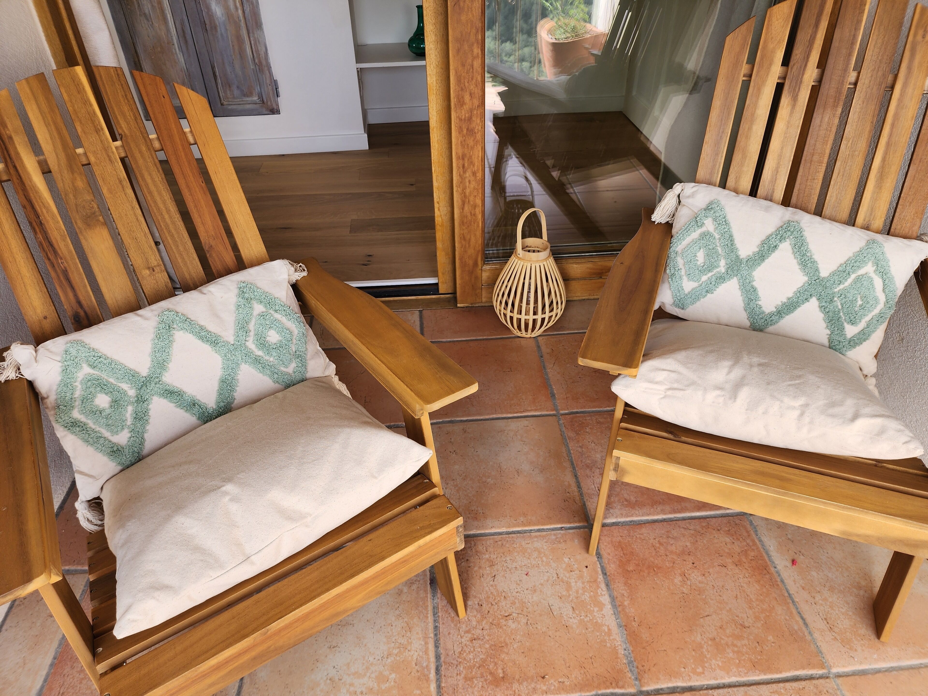 Two wooden chairs with cushions, a woven lantern, and a view of a room with a window and a potted plant.