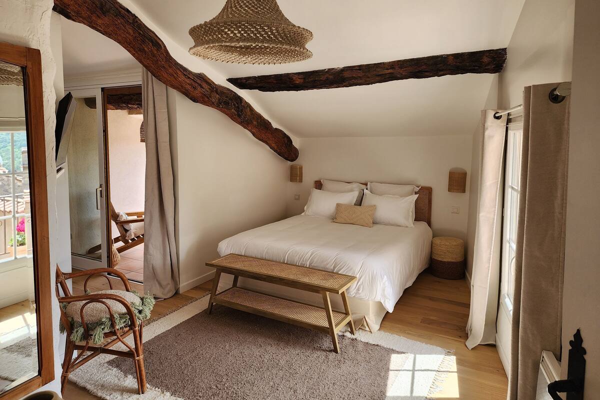 A bedroom with a large bed, a wooden bench, a woven light fixture, and exposed wooden beams.