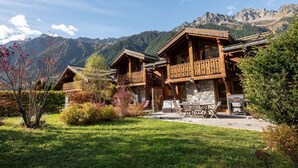 Un bâtiment de style chalet, doté de balcons en bois et de fondations en pierre, entouré d’un jardin bien entretenu et avec des montagnes en toile de fond.