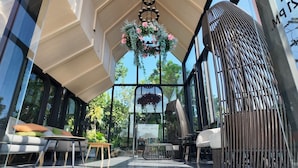 Ein Sitzbereich im Freien mit modernem Design, mit einer hängenden Blumenkomposition, einer Metallkonstruktion für einen Grill und Blick auf die umliegende Bepflanzung.