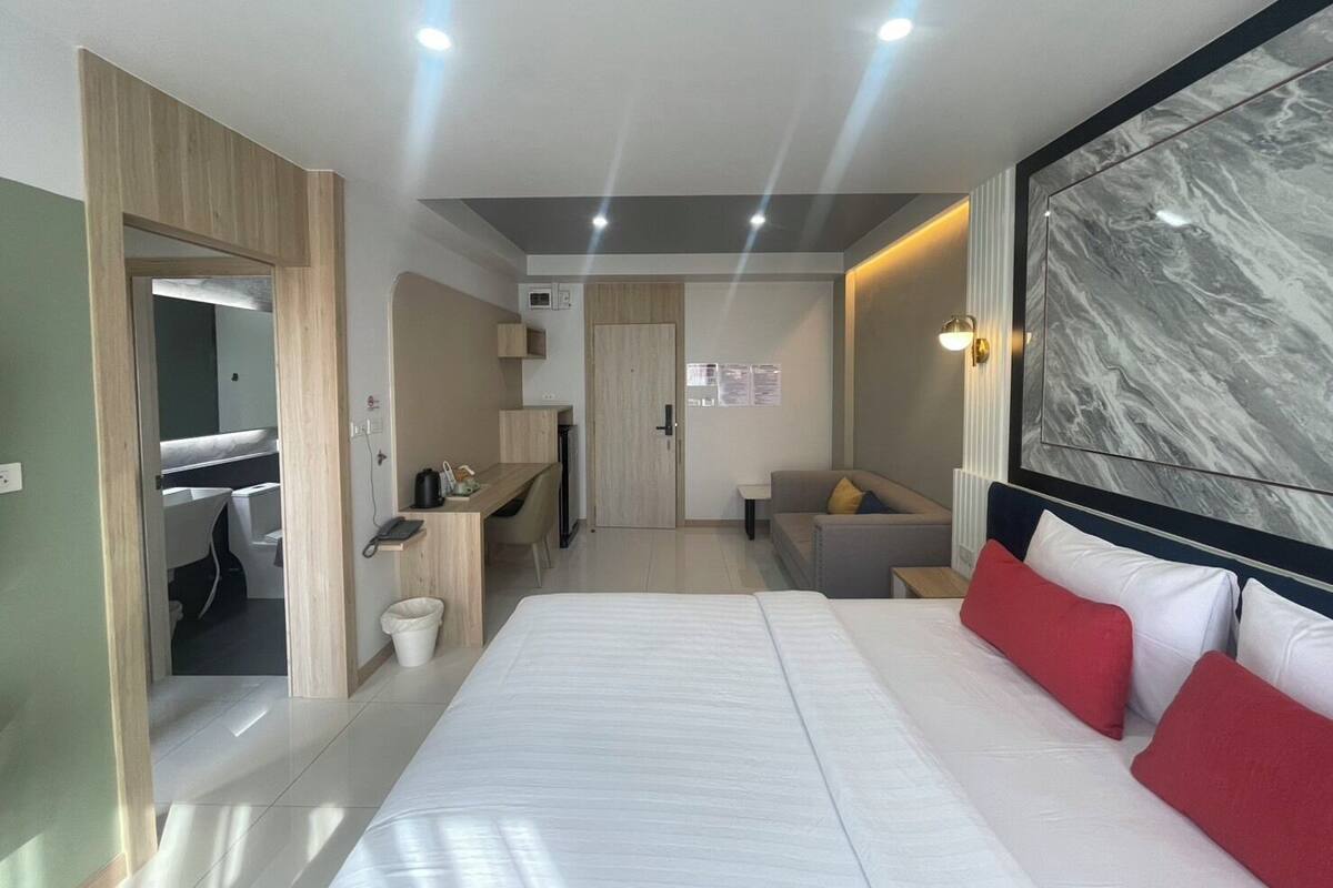 Une chambre d’hôtel moderne dotée d’un grand lit, d’un coin salon avec un canapé et des fauteuils, d’un petit bureau avec une chaise et d’une salle de bain visible à travers une porte ouverte.