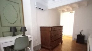 A room with a white desk, two lamps, a green abstract painting, a wooden dresser, and a tiled floor.