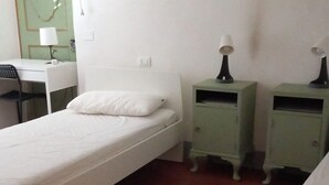 A single bed with a white mattress and pillows, a desk with a lamp, two green bedside tables with lamps, and a red carpeted floor.