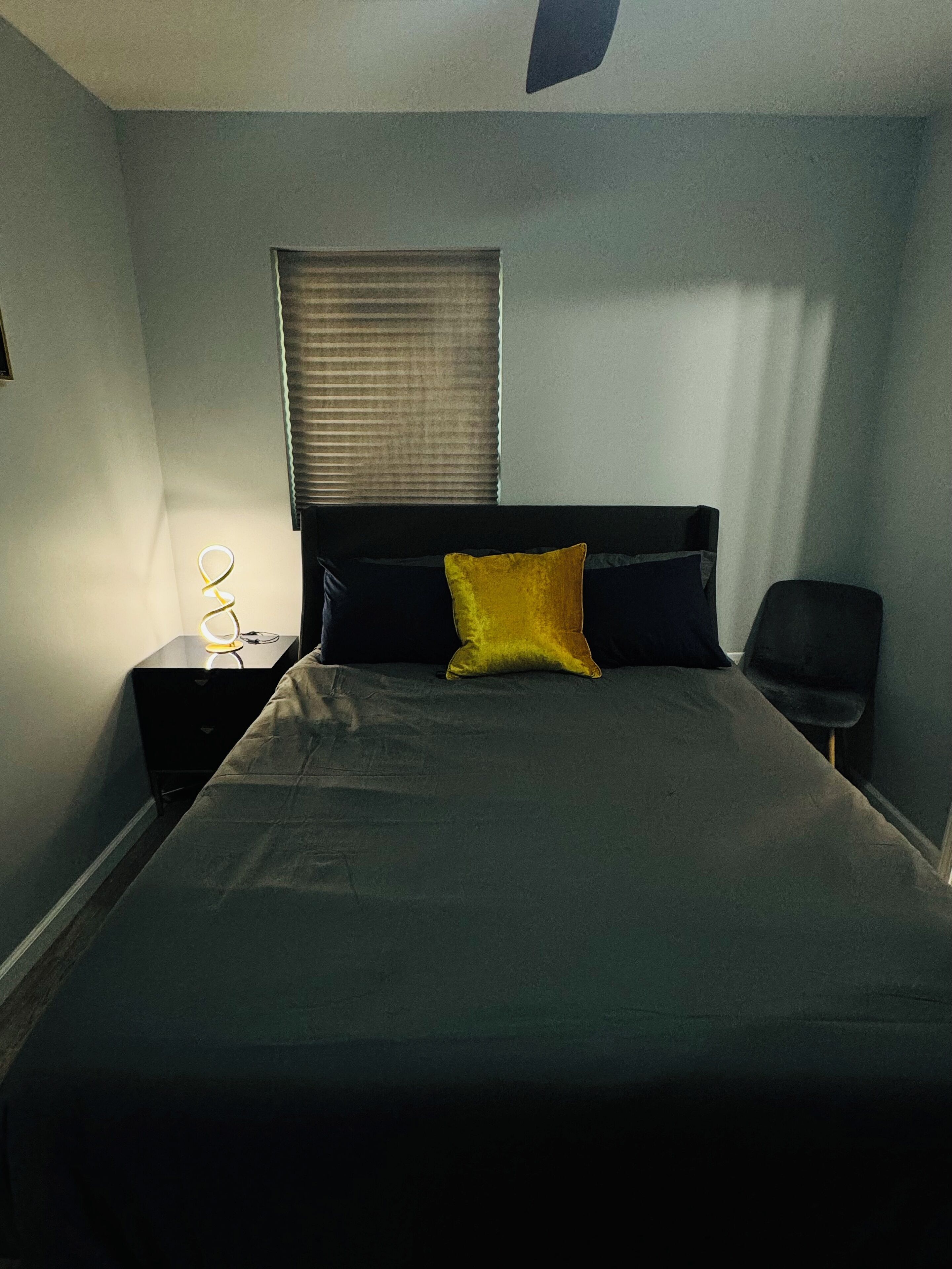 A bedroom with a bed, a nightstand, a lamp, a chair, and a window with blinds.