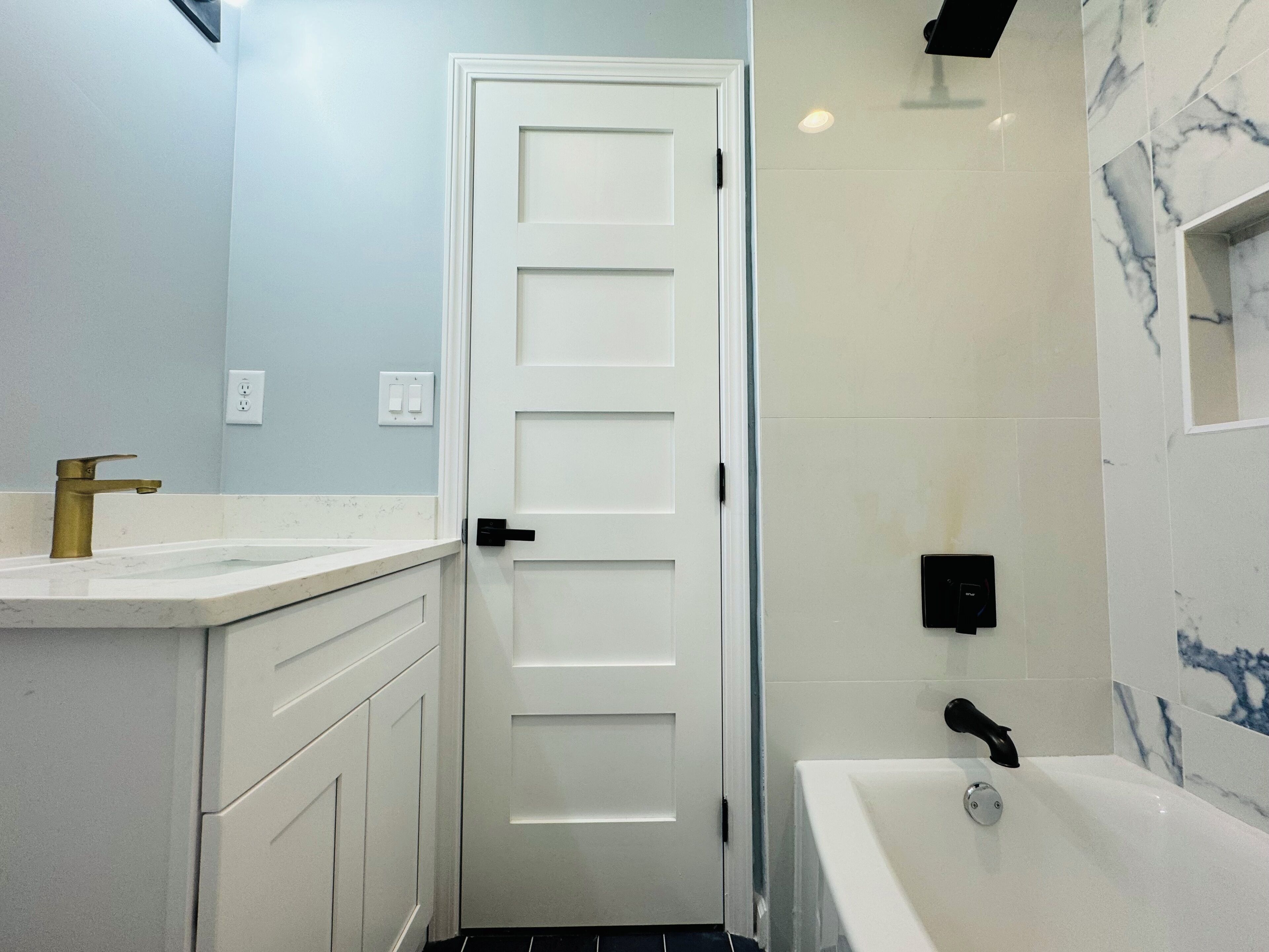 A bathroom with a white door, a sink with a gold faucet, a bathtub, and marble-patterned walls.