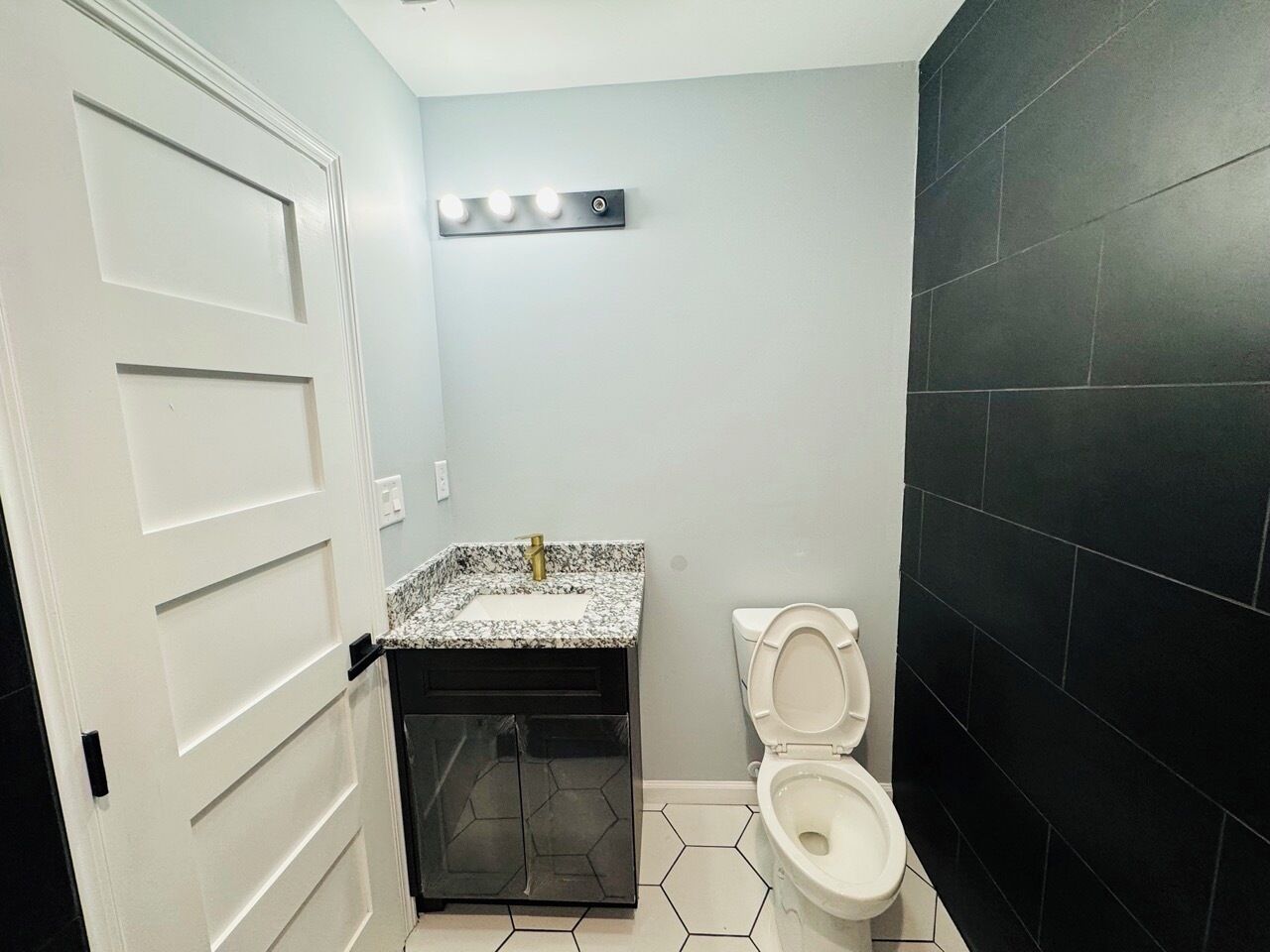 A bathroom with a white door, a sink with a marble countertop, a toilet, and a black tiled wall.