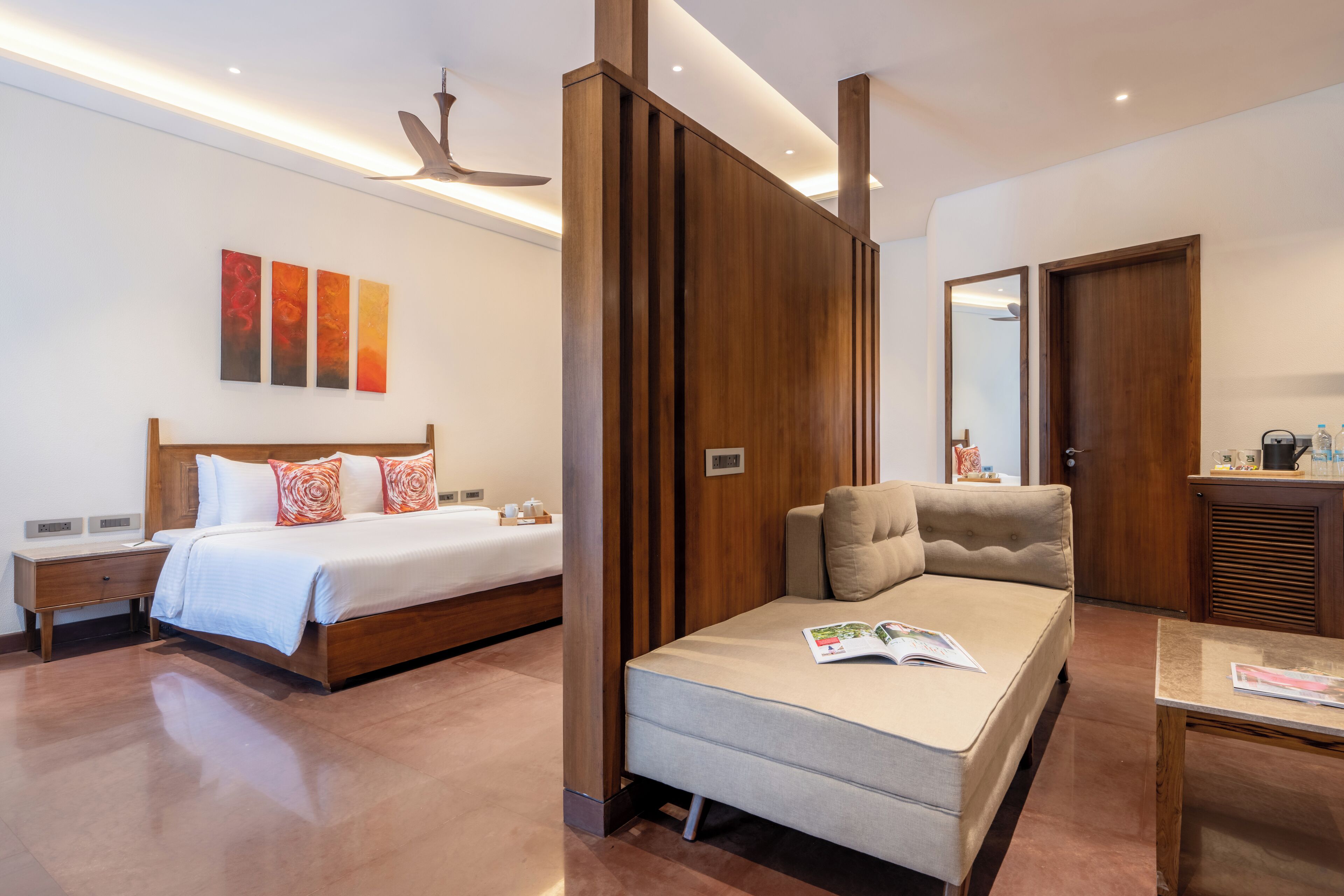 A modern hotel room with a large bed, a sofa, a desk, and a bathroom visible through a wooden partition.