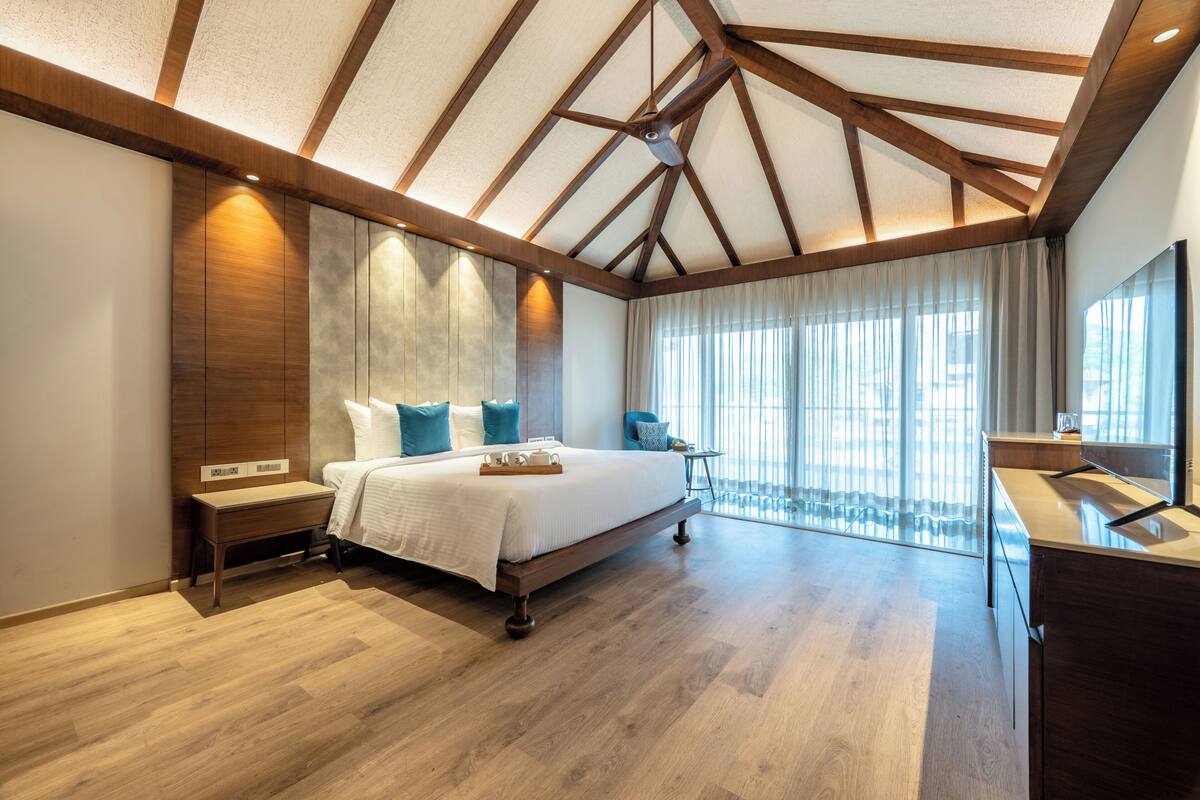 A spacious bedroom with a large bed, wooden beams on the ceiling, and a view of the outdoors through large windows.