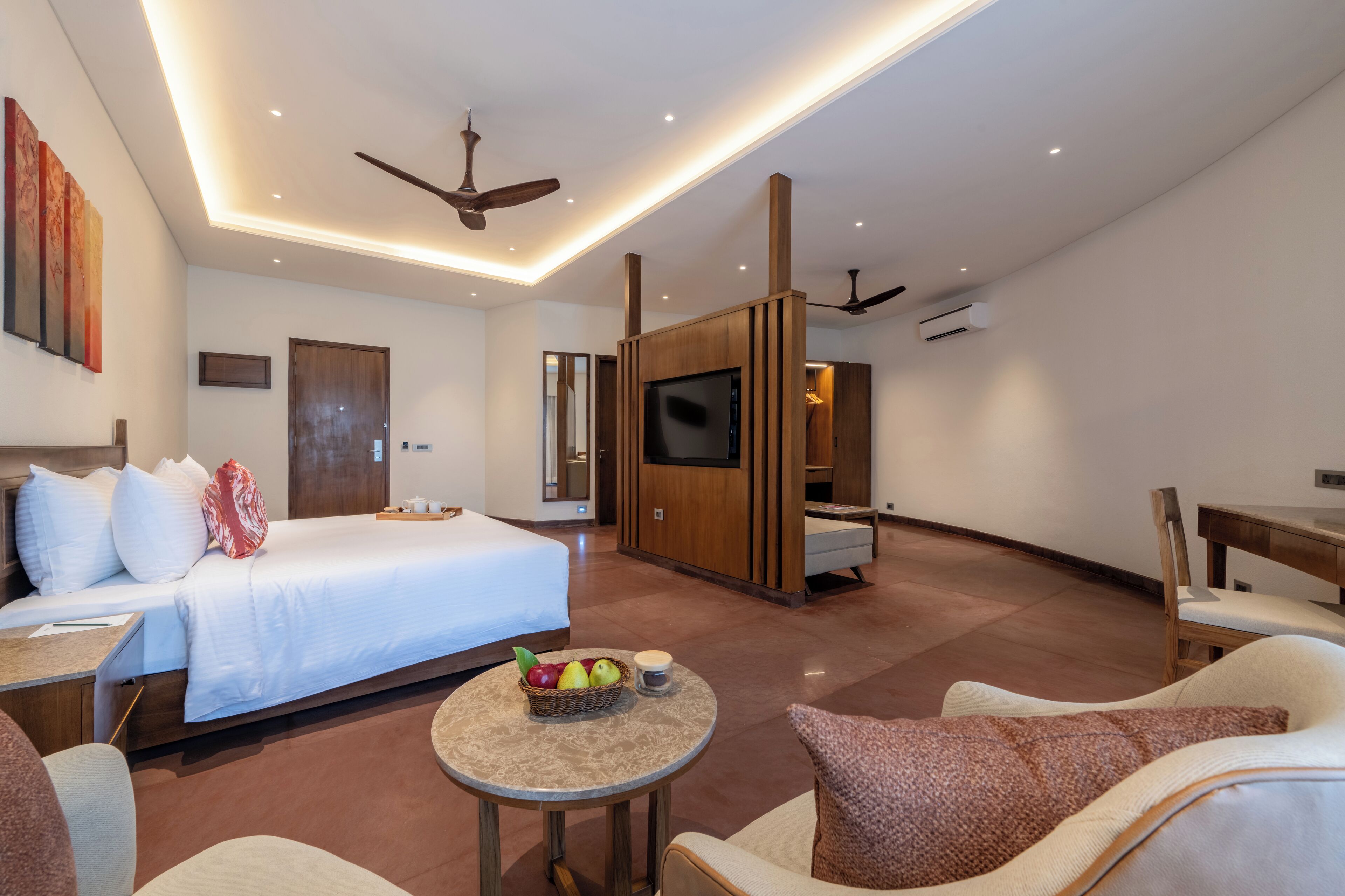A modern hotel room with a bed, seating area, TV, and a small table with fruit.