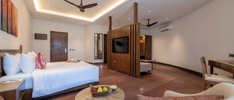 A modern hotel room with a bed, seating area, TV, and a small table with fruit.