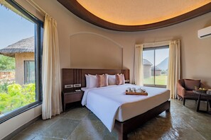 A hotel room with a large bed, a nightstand, a chair, a table, and a view of greenery and a thatched-roof structure outside.