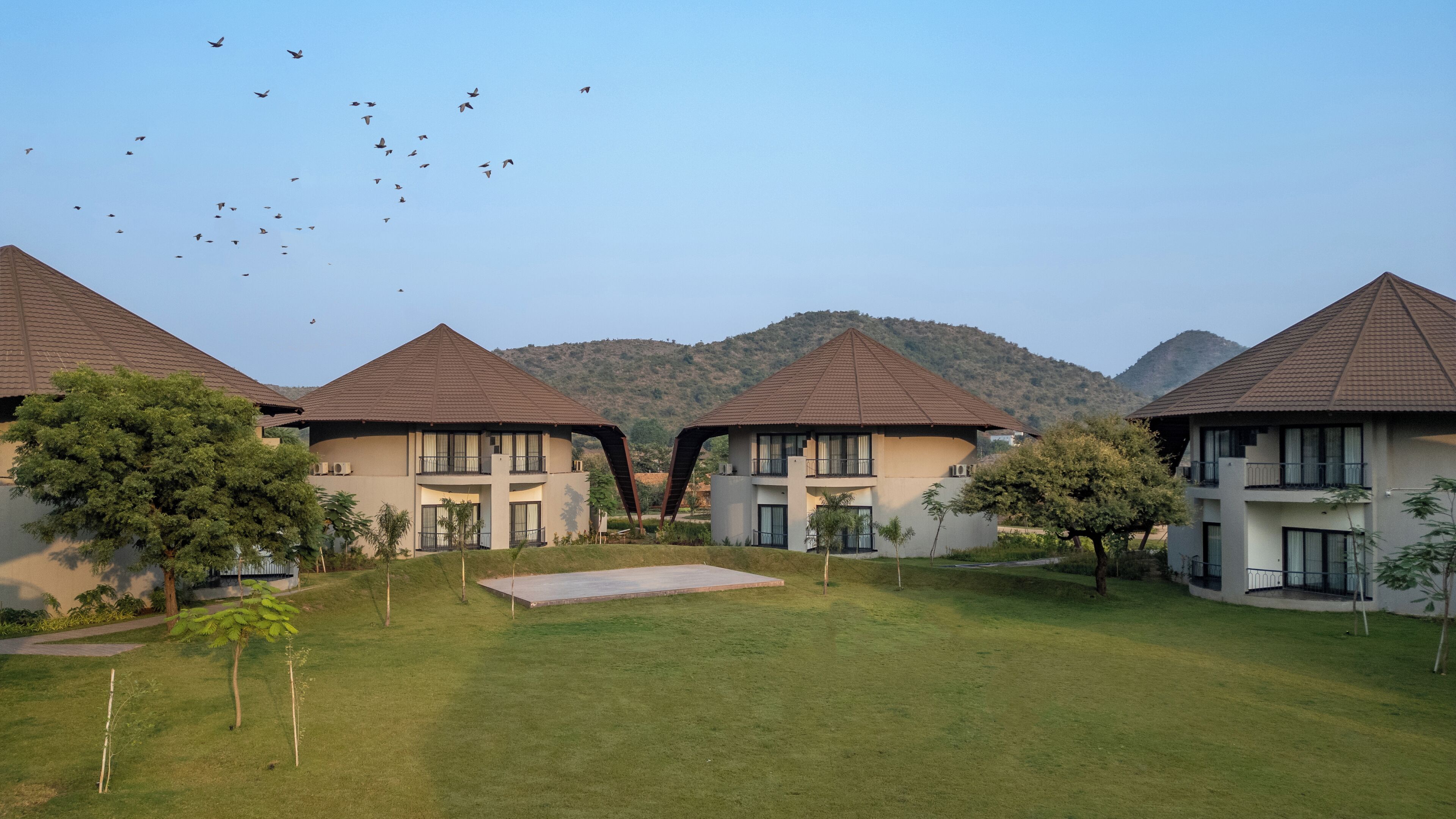 Un resort con varias edificaciones de dos pisos, cada una con techo marrón y ventanas amplias. En primer plano, un área con césped, algunos árboles y un cielo despejado al fondo.