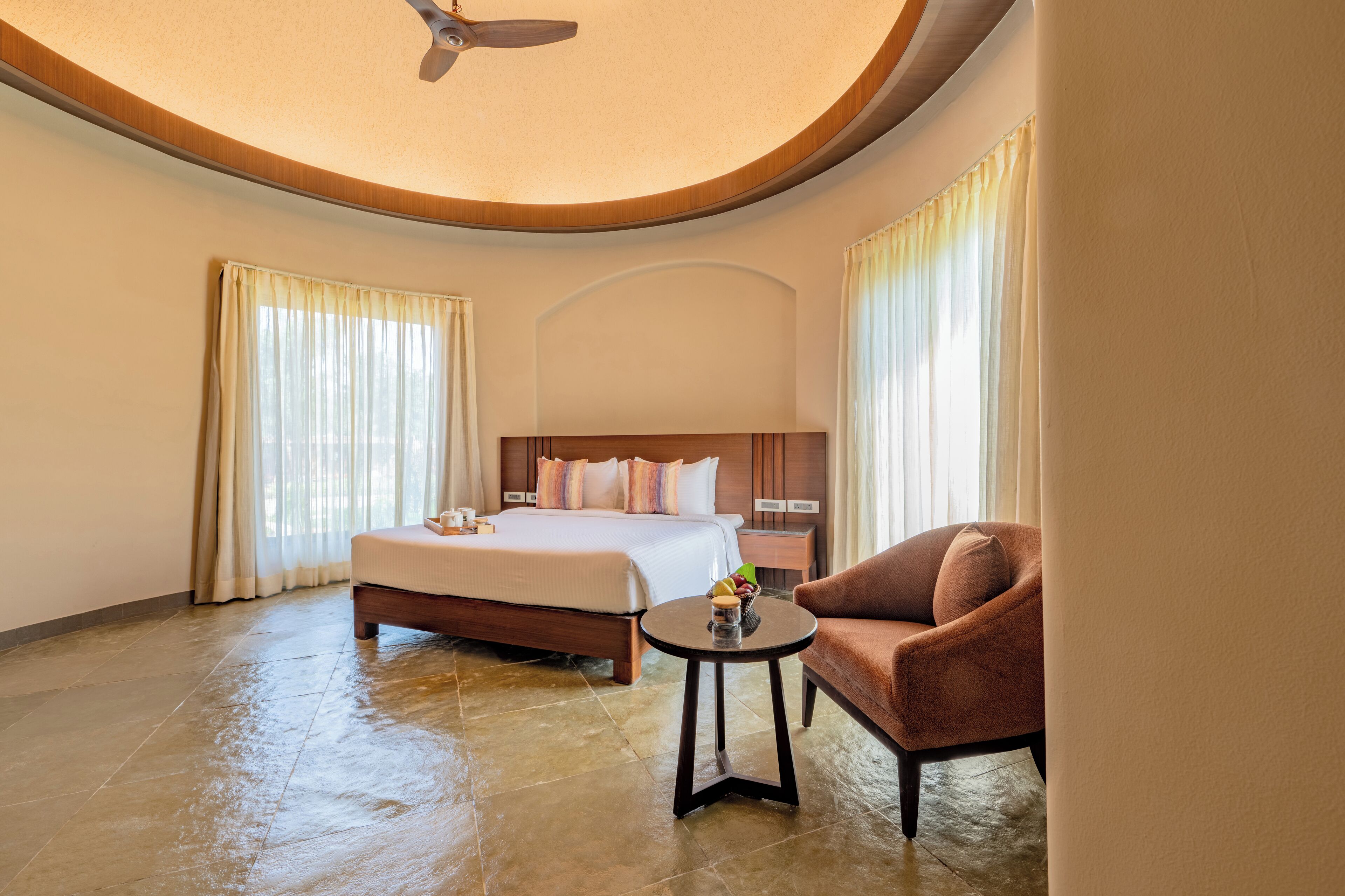 A hotel room with a large bed, a round table, a brown armchair, and a ceiling fan.