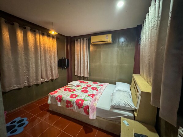Habitación de hotel con cama, cortinas, unidad de aire acondicionado, suelo de baldosas y una mesita con un control remoto.