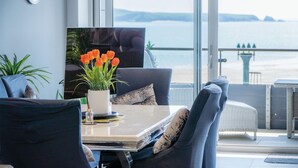 Ein modernes Wohnzimmer mit Meerblick, ausgestattet mit einem Esstisch mit einer Vase orangener Blumen, einer Couch und einem Fernseher.