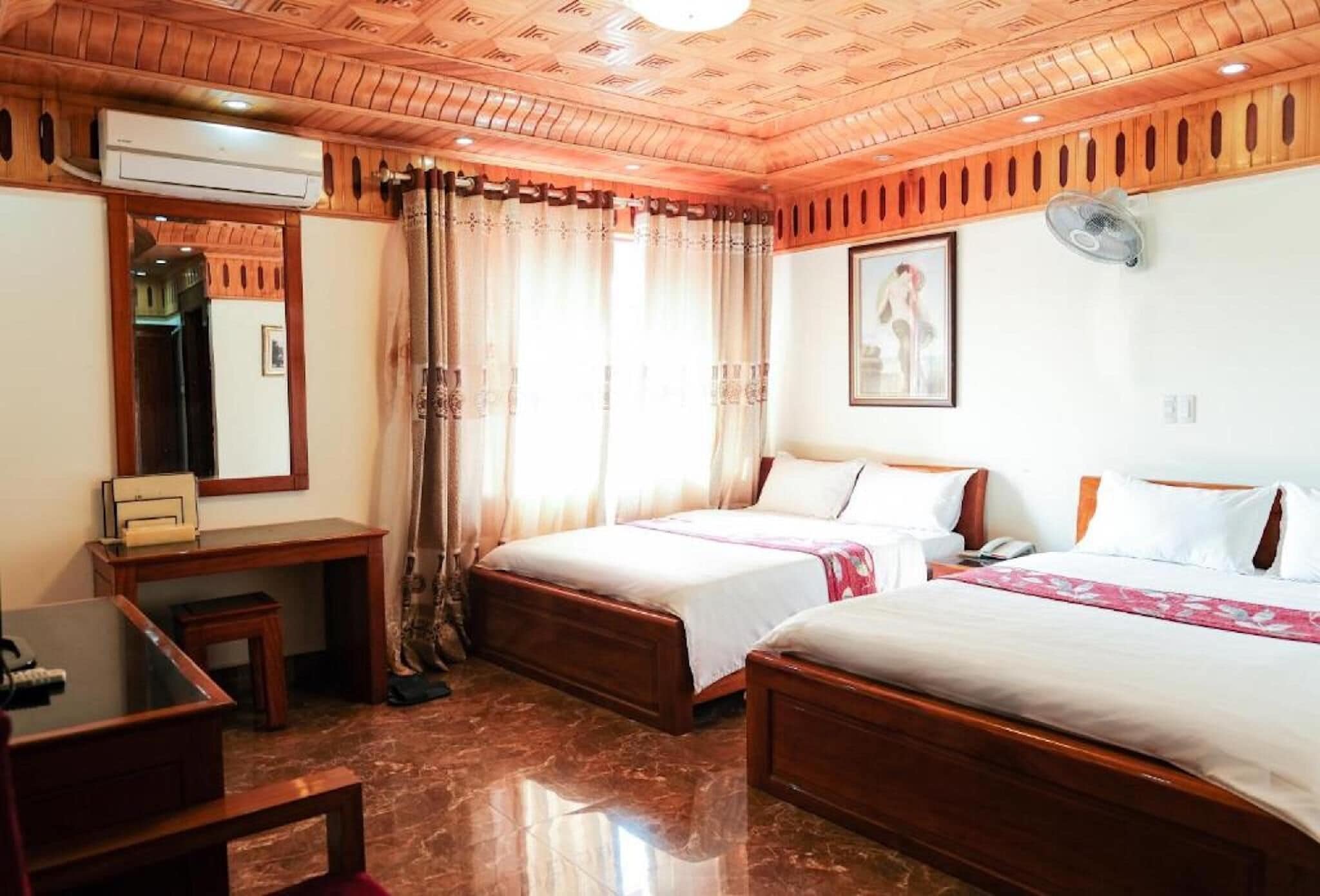 A hotel room with two beds, a desk, a chair, an air conditioning unit, and a ceiling fan.