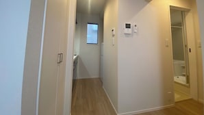 Ein schmaler Korridor mit Holzboden, weißen Wänden und einem kleinen Fenster. Es sind zwei an der Wand befestigte Geräte angebracht, möglicherweise Thermostate oder Gegensprechanlagen.