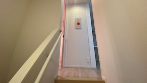 Eine enge Treppe mit Handlauf, einer Lampe und einem roten Notrufknopf an der Wand.