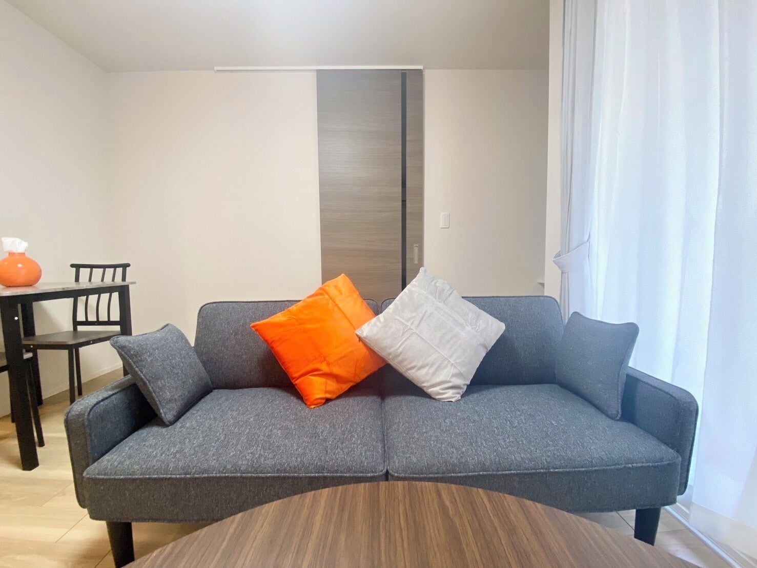 Una sala moderna con un sofá gris, dos almohadas, una mesa de centro redonda de madera y un comedor con mesa y sillas.