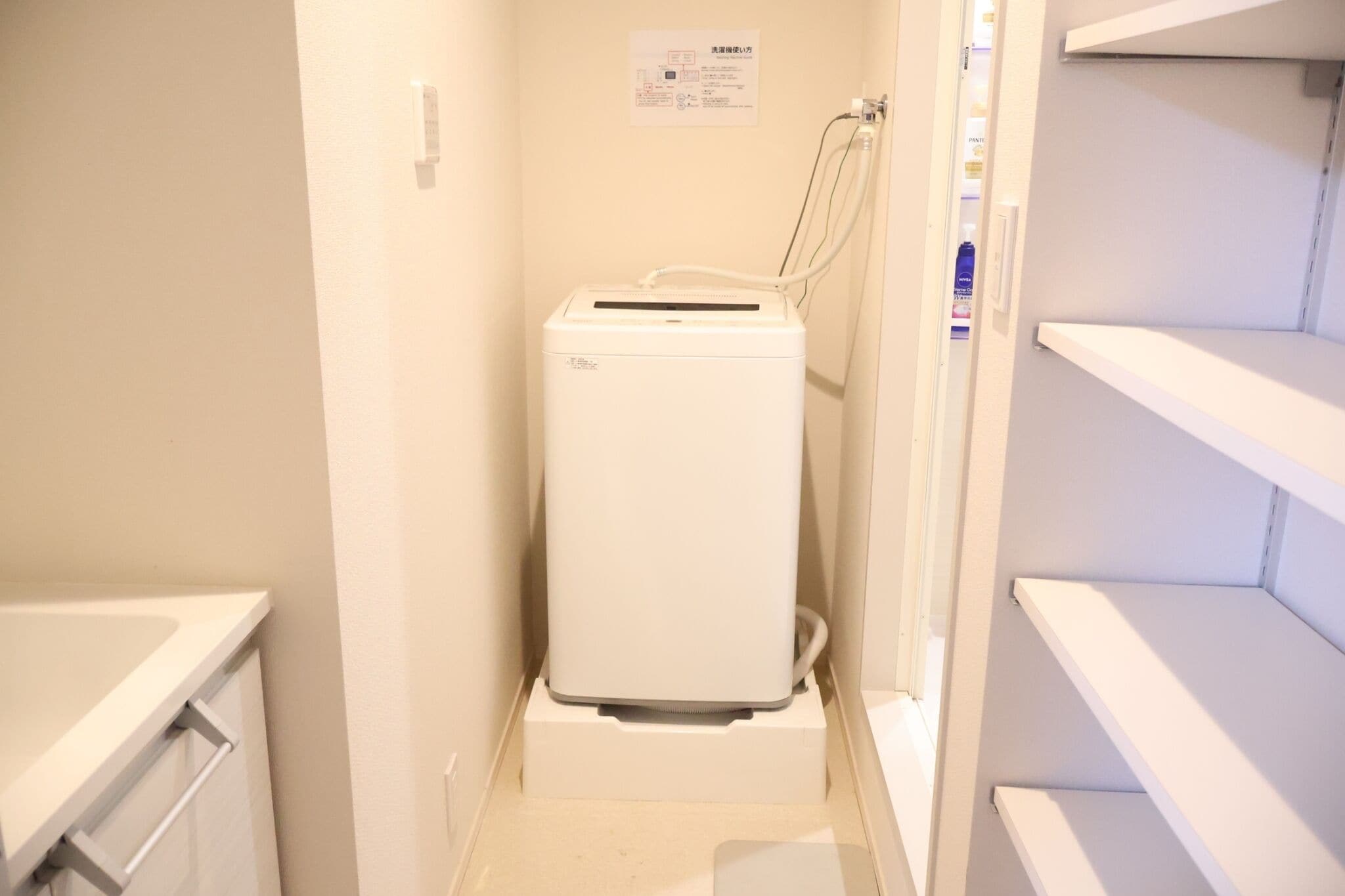 Een kleine, witte wasmachine in een smalle gang met witte muren en planken.