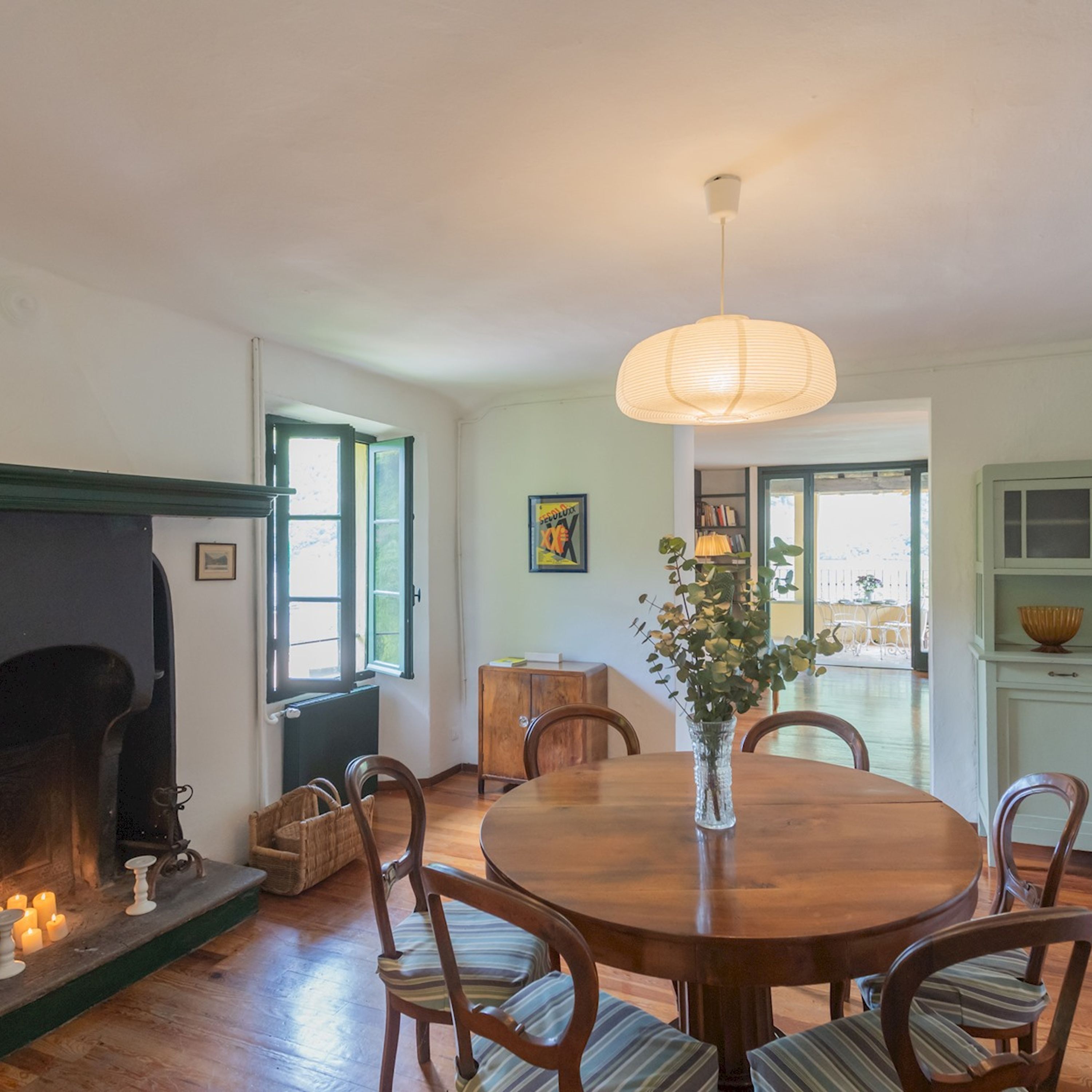 A dining area with a round wooden table, chairs, a vase with flowers, and a hanging light fixture.