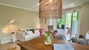 A modern living room with a wooden dining table, a sofa with patterned cushions, and a hanging woven light fixture. - Hundefreundliche Neubauwohnung Direkt am Wasser! (Eilsum)