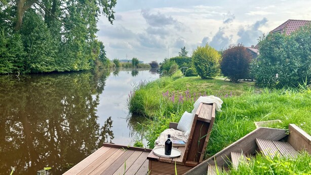 A wooden deck with a lounge chair and a small table, overlooking a calm river and lush greenery. - Hundefreundliche Neubauwohnung Direkt am Wasser! (Eilsum)