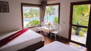 A hotel room with two beds, a small table with chairs, and large windows offering a view of a swimming pool and greenery. - Tràng An Hometown Homestay (Ninh Binh)