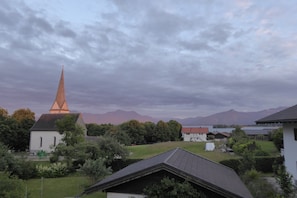 Eine Kirche mit einem hohen Turm, umgeben von Bäumen und Häusern, mit Bergen im Hintergrund.