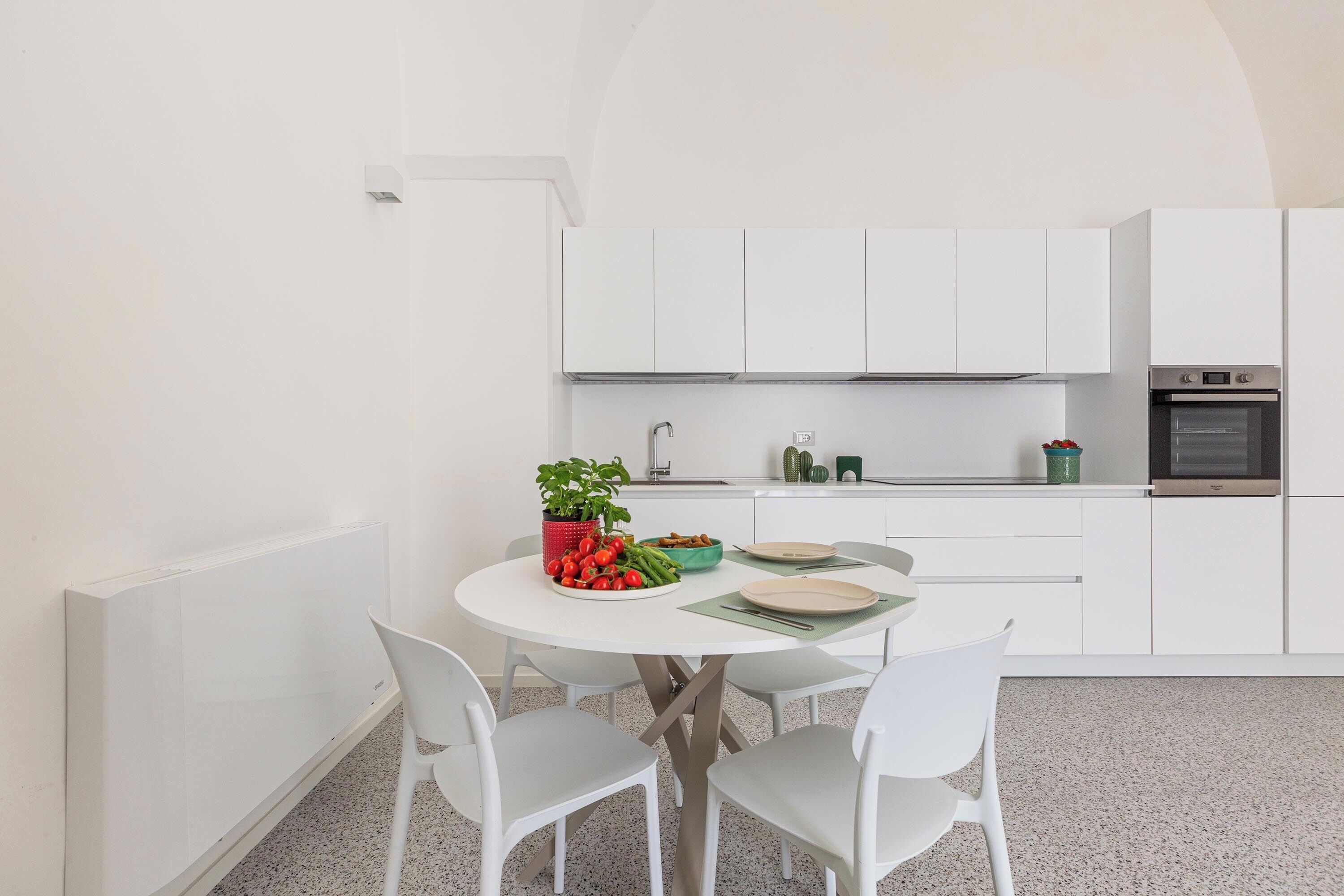 A modern kitchen with white cabinets, a dining table with chairs, and a small countertop with a plant and vegetables.