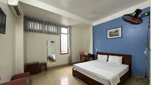Un dormitorio con una cama grande, un escritorio, una silla, un ventilador de pie y una ventana con cortinas.